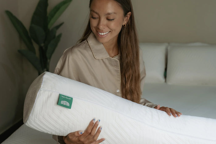 A woman holding a Lazy Lowie latex bolster (guling) on a bed